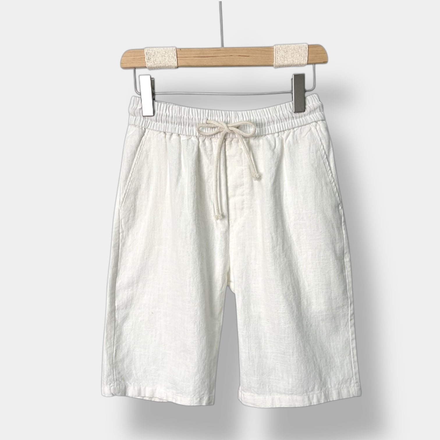White linen shorts with a drawstring on a wooden hanger against a white background