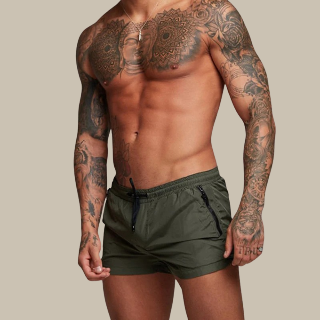 Man wearing green swim shorts with tattoos on arms and chest against a beige background