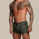 Man wearing green swim shorts with tattoos on arms and chest against a beige background