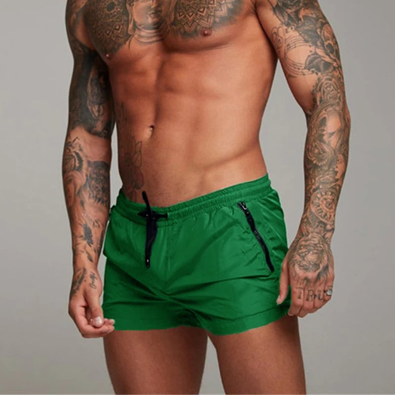 Man wearing green swim shorts with tattoos on arms and chest against a gray background